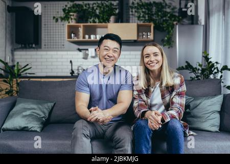 Liebevolles multirassisches Paar, Mann und Frau, Porträt einer jungen Familie, Blick auf die Kamera und lächelnd, sitzend auf dem Sofa im Wohnzimmer zu Hause Stockfoto