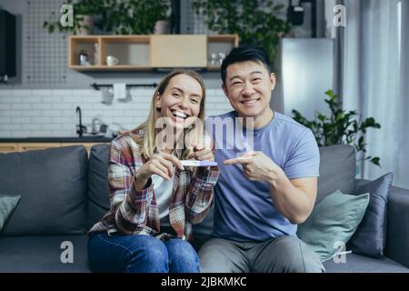 Junge glückliche asiatische Familie und Frau, die die Kamera anschaut und lächelt, einen positiven Schwangerschaftstest erhält und zu Hause auf der Couch sitzt Stockfoto