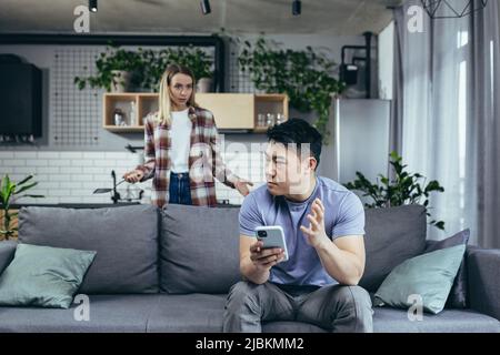 Zu Hause streitende Paare, Familienkrise, ein asiatischer Mann, der am Telefon auf der Couch sitzt, eine zornige Frau, die streitet und schreit Stockfoto