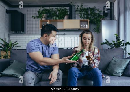 Alkoholikerin, die zu Hause harten Alkohol trinkt, Ehemann frustriert und verärgert, Konflikt in der jungen Familie um Alkohol Stockfoto