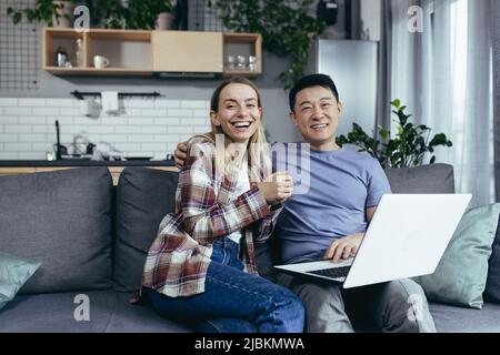 Junges Paar Asiatischer Mann und Frau zu Hause sitzen auf dem Sofa zusammen mit Laptop, glücklich und lächelnd multirassische Familie Blick auf die Kamera Stockfoto
