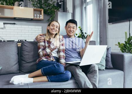 Junges Paar Asiatischer Mann und Frau zu Hause sitzen auf dem Sofa zusammen mit Laptop, glücklich und lächelnd multirassische Familie Blick auf die Kamera Stockfoto