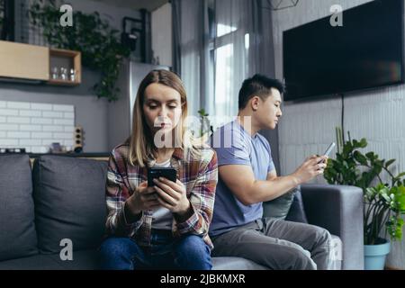Junge, multirassische Familie, streitsüchtig auf der Couch im Zimmer sitzend, Mann und Frau am Telefon, drehte sich in verschiedene Richtungen Stockfoto