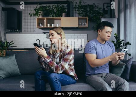 Junge, multirassische Familie, streitsüchtig auf der Couch im Zimmer sitzend, Mann und Frau am Telefon, drehte sich in verschiedene Richtungen Stockfoto