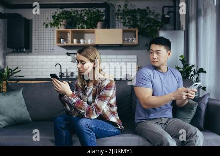 Junge, multirassische Familie, streitsüchtig auf der Couch im Zimmer sitzend, Mann und Frau am Telefon, drehte sich in verschiedene Richtungen Stockfoto