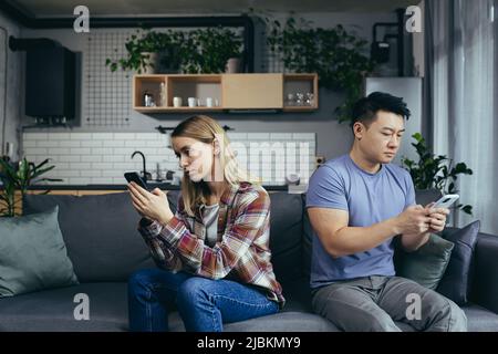 Junge, multirassische Familie, streitsüchtig auf der Couch im Zimmer sitzend, Mann und Frau am Telefon, drehte sich in verschiedene Richtungen Stockfoto
