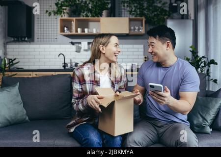 Das junge glückliche Paar erhielt ein Paket, glücklich und zufrieden mit dem Einkaufen im Online-Shop, Mann und Frau zu Hause sitzen auf dem Sofa Stockfoto