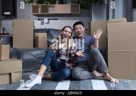 Junge, multirassische Familie, Mann und Frau, die zwischen Kartons auf dem Boden sitzen, auf die Kamera schauen und lächelnde, winkende Hände zum Gruß bewegen Stockfoto