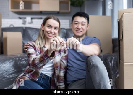 Porträt eines jungen Paares, eines Mannes und einer Frau, die auf die Kamera blicken und lächeln und die Schlüssel für eine neue Wohnung halten, eine multirassische Familie, die auf dem Bett sitzt Stockfoto