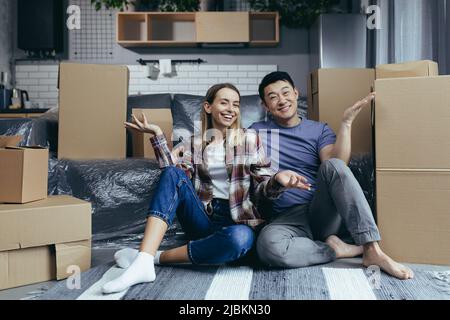 Junge, multirassische Familie, Mann und Frau, die zwischen Kartons auf dem Boden sitzen, auf die Kamera schauen und lächelnde, winkende Hände zum Gruß bewegen Stockfoto