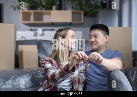 Nahaufnahme einer jungen Familie in einer neuen Wohnung, die die Schlüssel zum Haus hält, ein multirassisches Paar, das im Wohnzimmer zwischen dem Karton sitzt Stockfoto