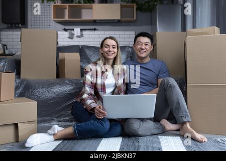 Junge glückliche multirassische Familie in einer neuen Wohnung mit einem Laptop, Asiatin und Frau, die auf die Kamera schauen und lächeln, mit Freunden online chatten, zeigen Stockfoto