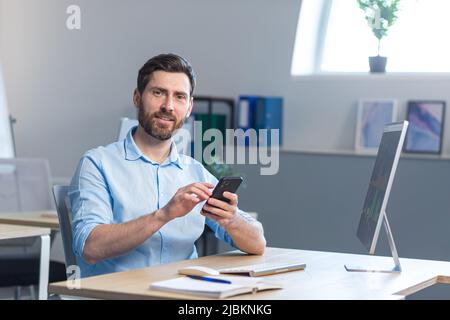 Erfolgreicher Geschäftsmann mit Bart blickt auf die Kamera und lächelt, nutzt ein Mobiltelefon, während er an einem Schreibtisch im Büro sitzt Stockfoto