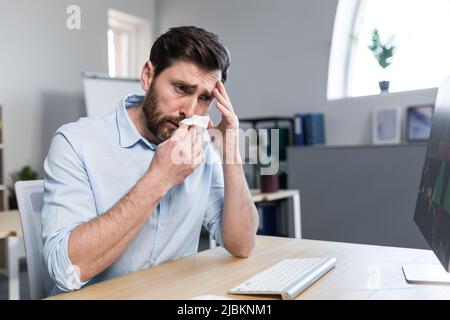 Kranker und müder junger Mann bei der Arbeit. Büroangestellter, Manager, Freiberufler, der an einem Schreibtisch im Büro sitzt, fühlt sich nicht gut an, wischt sich die Nase ab, hat ein laufendes n Stockfoto