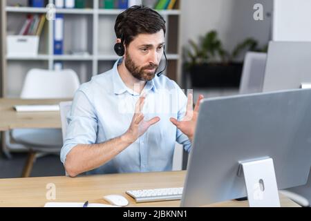 Ein junger Mann, ein Berater, ein Büroangestellter, ein Manager, der bei einem Videoanruf mit Kopfhörern spricht, seine Arme schwenkt und etwas erklärt. Sitzen in einem kostenlosen Stockfoto