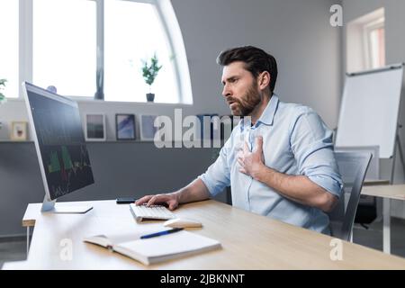 Kranker Geschäftsmann, der im Büro arbeitete, machte sich Sorgen und hielt seine Hand an seine Brust, schwere Herzschmerzen Stockfoto