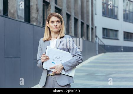 Porträt einer Geschäftsfrau, in grauem Anzug, außerhalb des Büros, einer Geschäftsfrau mit Laptop und Papierdokumenten Stockfoto