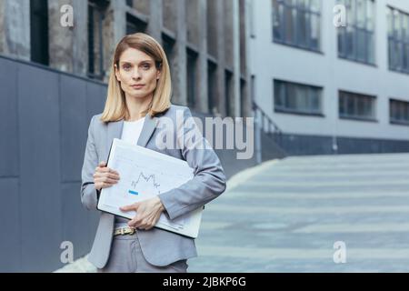 Porträt einer Geschäftsfrau, in grauem Anzug, außerhalb des Büros, einer Geschäftsfrau mit Laptop und Papierdokumenten Stockfoto