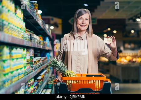 Porträt einer schönen älteren Frau in einem Geschäft, Supermarkt, mit einer Kreditkarte in den Händen. Steht zwischen den Regalen eines Ladens, Supermarkt w Stockfoto