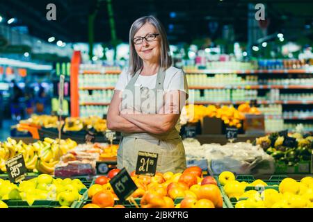 Porträt einer älteren grauhaarigen Frau eines Supermarktarbeiters, Arbeiter breitet Obst mit gekreuzten Armen lächelt und schaut auf die Kamera Stockfoto