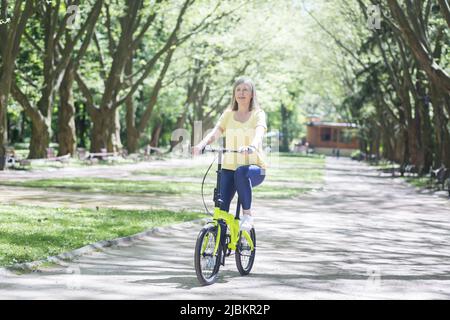 Eine aktive Rentnerin fährt in einem Sommerpark mit dem Fahrrad, eine ältere grauhaarige Rentnerin geht zu Fuß Stockfoto