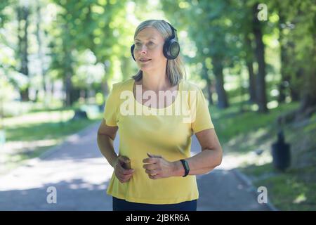 Ältere schöne Frau mit Kopfhörern, die im Park läuft, morgens joggt, aktiv ist, Musik hört. Fühlt sich gut an, lächelt Stockfoto