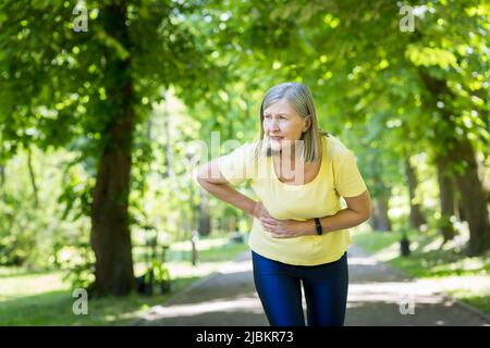 Ältere schöne Frau, die im Park läuft und Sport spielt. Ihr Magen begann zu Schmerzen, sie hielt sich an ihrem Bauch fest und beugte sich vor Schmerzen. Stockfoto