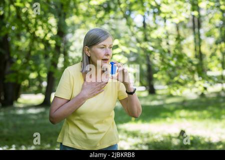 Saisonale Allergien. Eine ältere Frau im Park hat in der Natur Atembeschwerden, nutzt einen Inhalator. Niest und hustet in ein Taschentuch. Hat alle Stockfoto