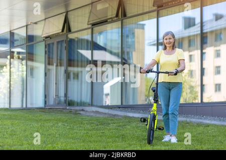 Porträt einer schönen älteren Frau. Stehen mit einem gelben Fahrrad in der Nähe des Bürozentrums. Fährt mit dem Fahrrad. Aktiver Lebensstil. Er schaut auf die Kamera Stockfoto