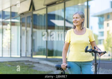 Eine aktive ältere Frau im Ruhestand geht mit dem Fahrrad vor ein modernes Gebäude Stockfoto