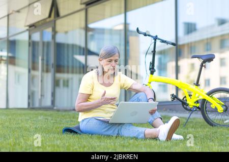 Ältere grauhaarige Rentnerin, die auf dem Gras sitzt und bei einem Videoanruf auf einem Laptop-Bildschirm spricht, am Sommertag mit einem Fahrrad Stockfoto