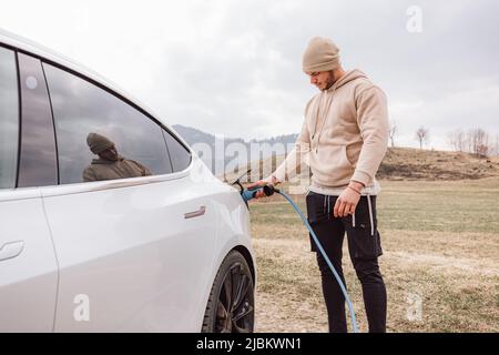 Männliche Hand, die ein Ladegerät in die Steckdose für Elektroautos steckt, im Hintergrund sichtbare Bergnatur Stockfoto