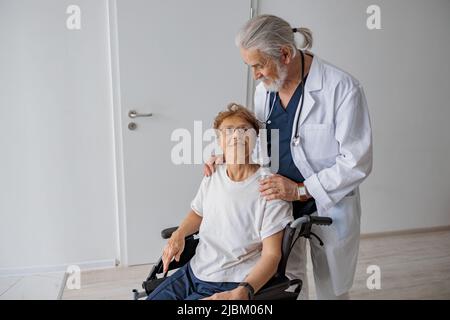 Professioneller männlicher Arzt, der weibliche Patientin auf dem Rollstuhl in der Medizinklinik trägt Stockfoto