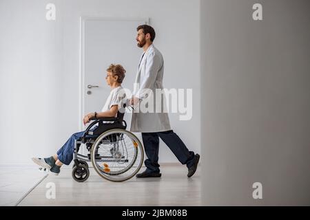 Professioneller männlicher Arzt, der weibliche Patientin auf dem Rollstuhl in der Medizinklinik trägt Stockfoto