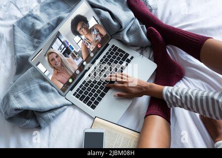 Junge Frau, die im Bett arbeitet, während sie zu Hause Videokonferenzen mit weiblichen Kollegen verschiedener Rassen führt Stockfoto