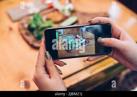 Food-Fotografie für soziale Netzwerke. Nahaufnahme der weiblichen Hände, die das Telefon mit dem Essen auf dem Bildschirm halten, und Aufnahme einer gesunden Mahlzeit. Stockfoto