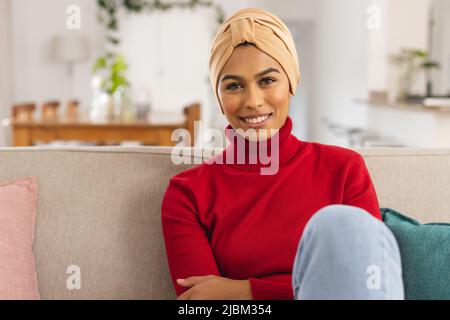 Porträt einer glücklichen jungen Birazialfrau mit Kopftuch, die zu Hause auf dem Sofa sitzt Stockfoto