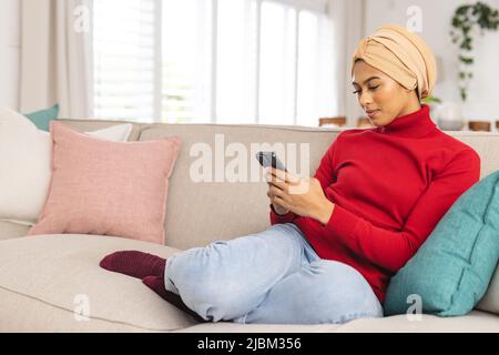 Biracial junge Frau im Kopftuch mit Smartphone, während sie auf dem Sofa zu Hause sitzen Stockfoto