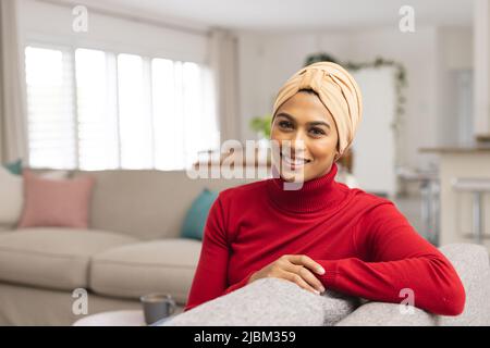 Porträt einer lächelnden jungen Biracial-Frau im Kopftuch, die zu Hause auf dem Sofa sitzt Stockfoto