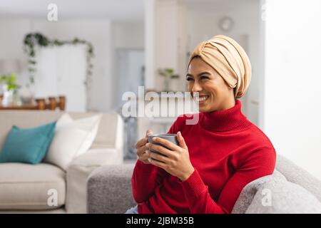 Glückliche junge Birazialfrau im Kopftuch beim Kaffee, während sie zu Hause auf dem Sofa sitzt Stockfoto