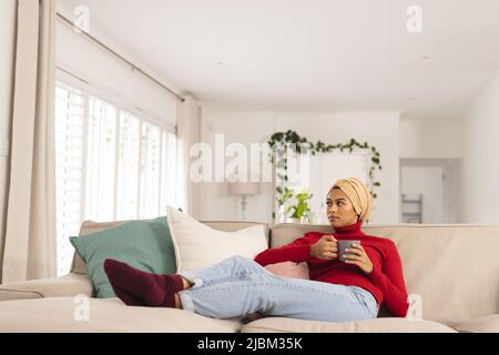 Nachdenkliche Biracial junge Frau im Kopftuch Kaffee trinken, während sie auf dem Sofa zu Hause sitzt Stockfoto