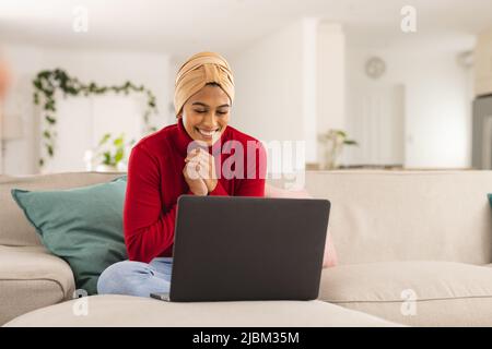 Aufgeregt biracial junge Frau im Kopftuch mit Laptop zu Hause Stockfoto