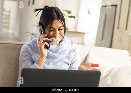 Glückliche junge Birazialfrau, die zu Hause mit einem Laptop auf einem Smartphone spricht Stockfoto