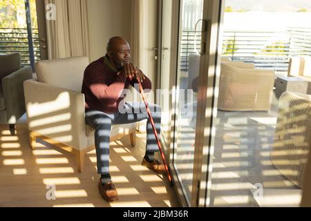 Kahlköpfiger afroamerikanischer Senioren mit Gehstock, der durch das Fenster schaute, während er auf einem Sessel saß Stockfoto