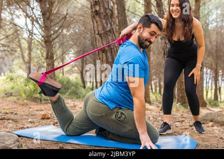 Hübsches Mädchen lehrt einen hübschen jungen Mann, Yoga-Strecken auf ihrer Matte mit einem rosa Band in einem Wald, der von Bäumen umgeben ist, zu machen. Stockfoto