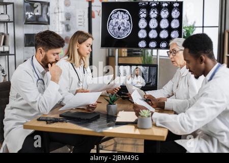 Multikulturelle Gruppe von vier medizinischen Forschern, die im Labor Online-Schulungen mit Monitor im Hintergrund absolvieren, der das MRT-Bild des Gehirns zeigt. Kollegen teilen Ideen, während sie die Liste der Patientenakten speichern. Stockfoto