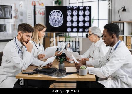 Multikulturelle Gruppe von vier medizinischen Forschern, die im Labor Online-Schulungen mit Monitor im Hintergrund absolvieren, der das MRT-Bild des Gehirns zeigt. Kollegen teilen Ideen, während sie die Liste der Patientenakten speichern. Stockfoto