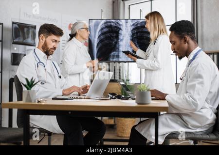 Team aus zwei mehrethnischen männlichen medizinischen Mitarbeitern, die auf drahtlosen Laptops tippen, während weibliche Kollegen Tomographieergebnisse diskutieren und Diagnosen stellen. Gesundheitswesen und modernes Technologiekonzept. Stockfoto