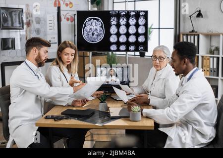 Multikulturelle Gruppe von vier medizinischen Forschern, die im Labor Online-Schulungen mit Monitor im Hintergrund absolvieren, der das MRT-Bild des Gehirns zeigt. Kollegen teilen Ideen, während sie die Liste der Patientenakten speichern. Stockfoto