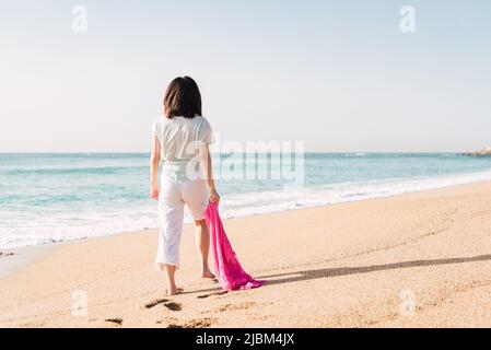 Rückansicht eines nicht erkennbaren Weibchens in weißer Kleidung und mit Schal, das bei sonnigem Wetter an der sandigen Küste nahe dem Meer spazieren geht Stockfoto